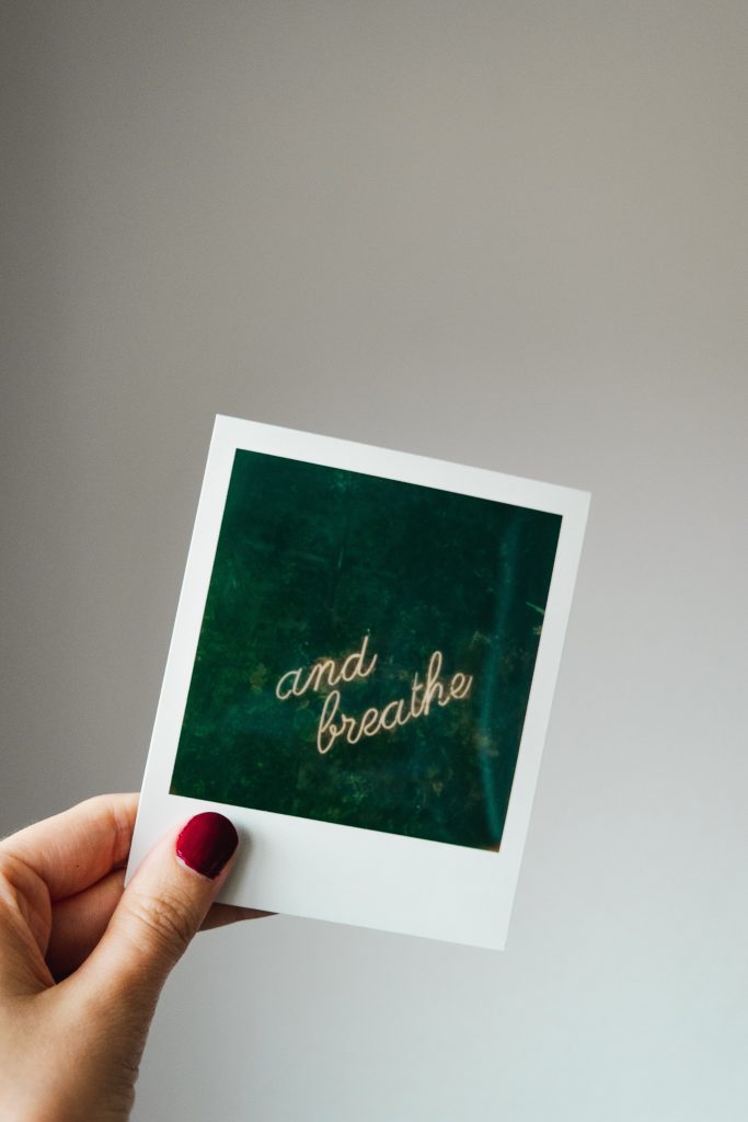 mano di donna con smalto rosso che regge polaroid con scritta a neon su fondo verde "and breath"