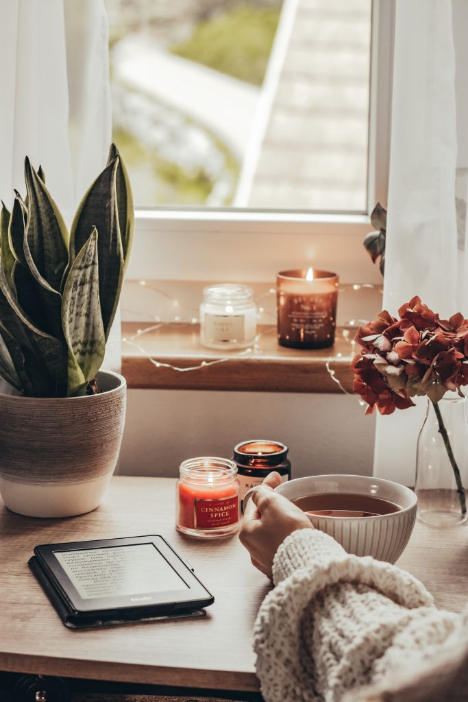 tavolo vicino finestra con candele, pianta, kindle, fiori secchi, tazza di te e mano di donna che regge la tazza