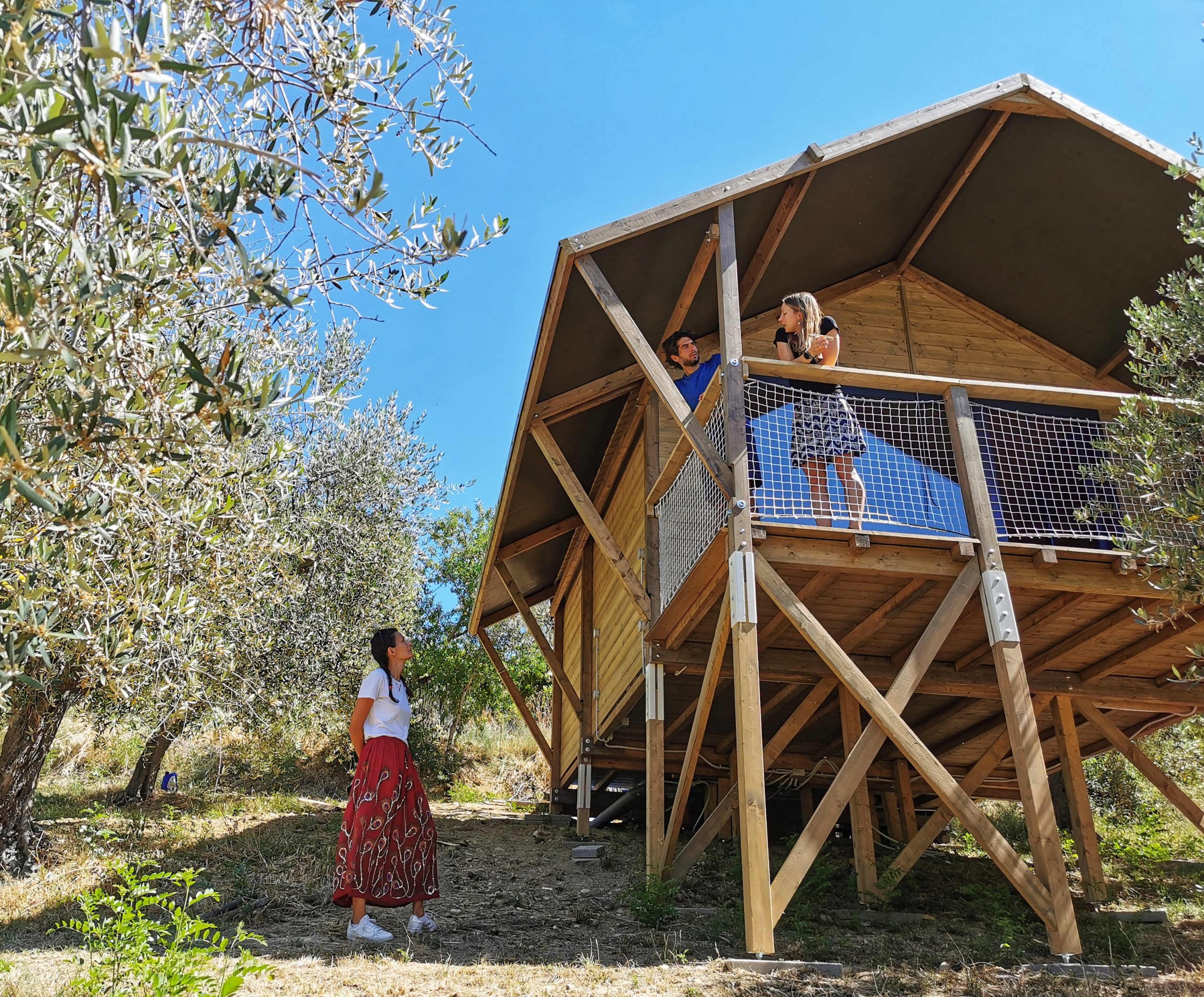 glamping in legno tra gli ulivi, due persone sul terrazzo e una donna con gonna rossa tra gli ulivi