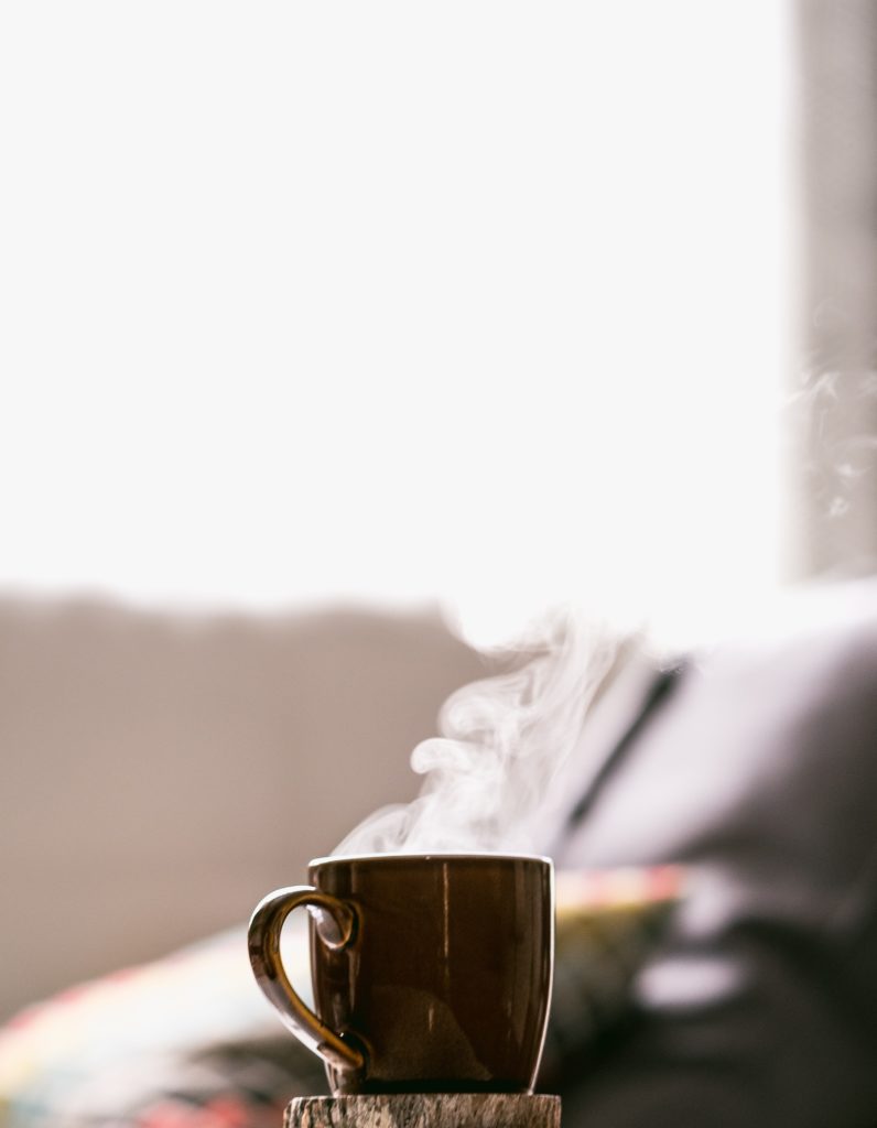 tazza di caffè fumante marrone appoggiata su un cerchio di legno, luce mattutina, letto nello sfondo
