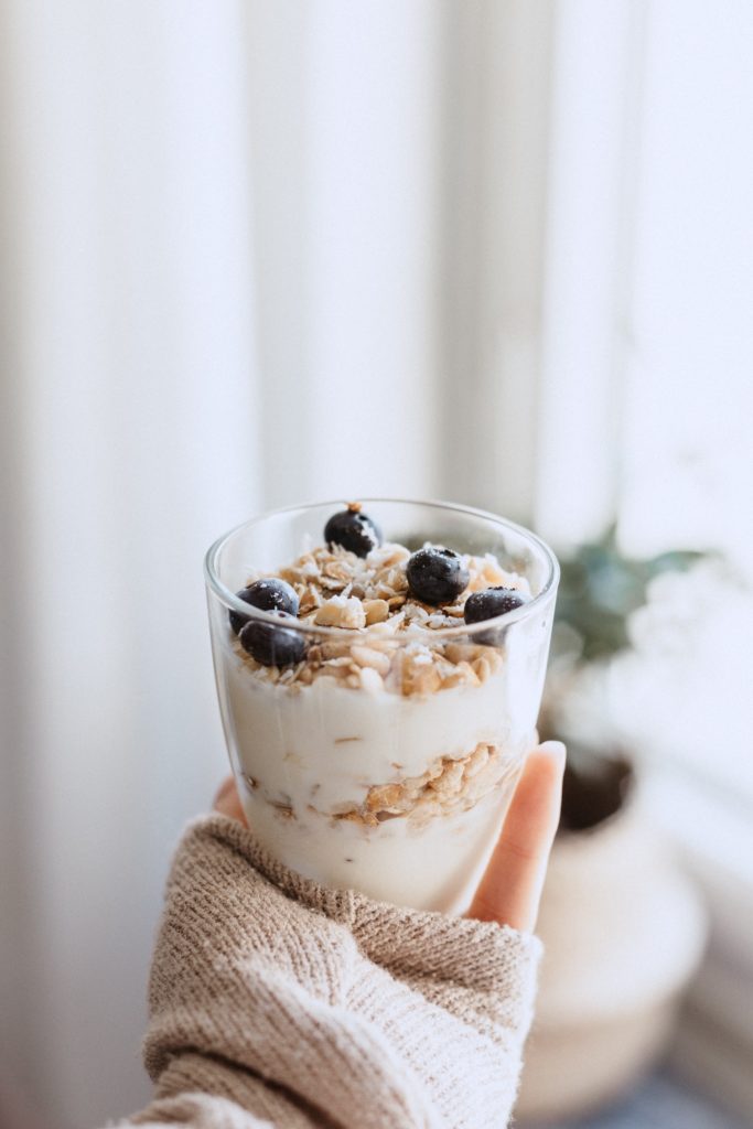 Mano di ragazza che tiene in mano un bicchiere con yogurt, avena e frutta. Sfondo sfocato